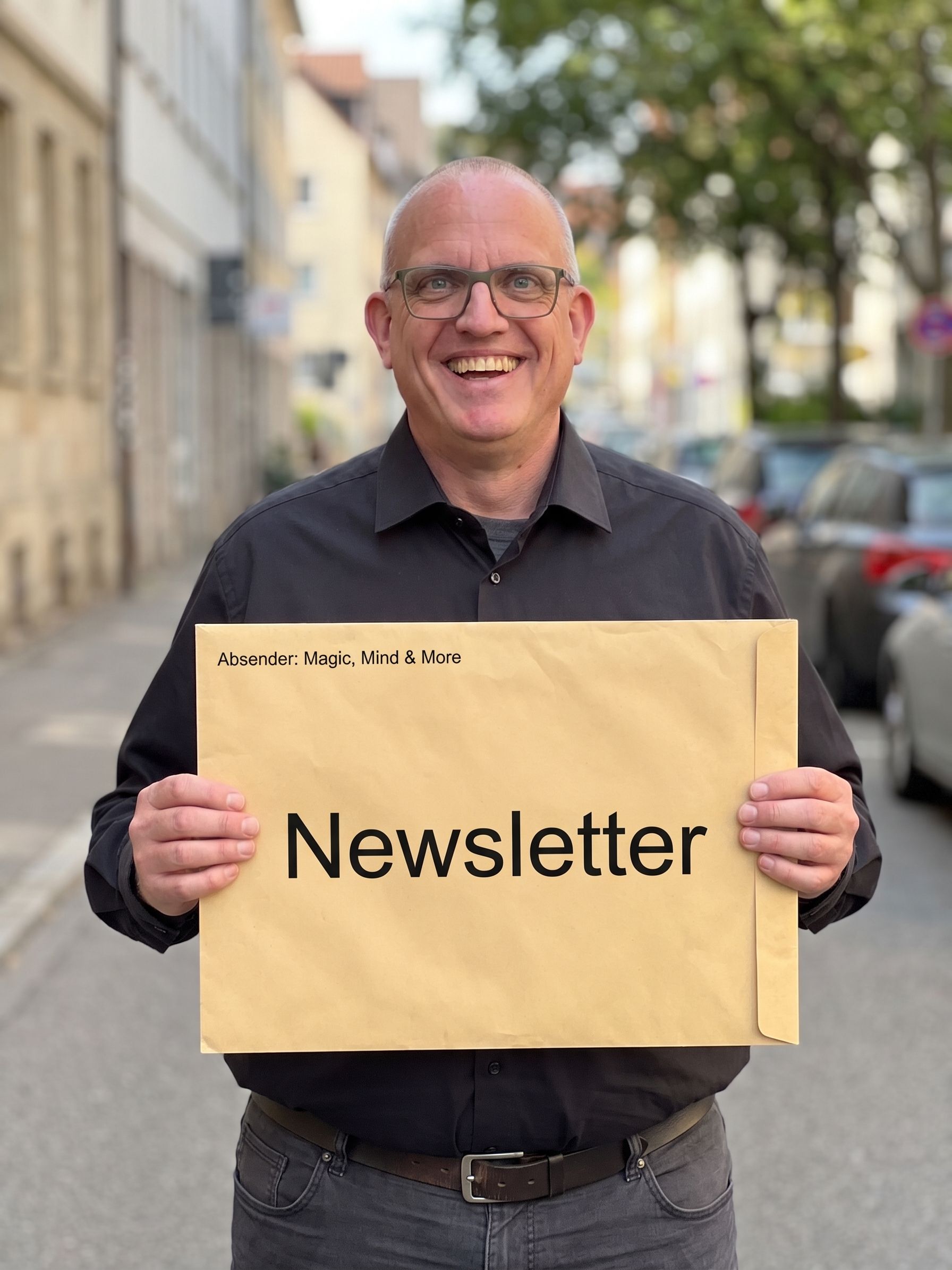 Joachim Jögel lachend auf der Straße, mit einem großen Umschlag mit der Aufschrift „Newsletter“ und „Absender: Magic, Mind & More“
