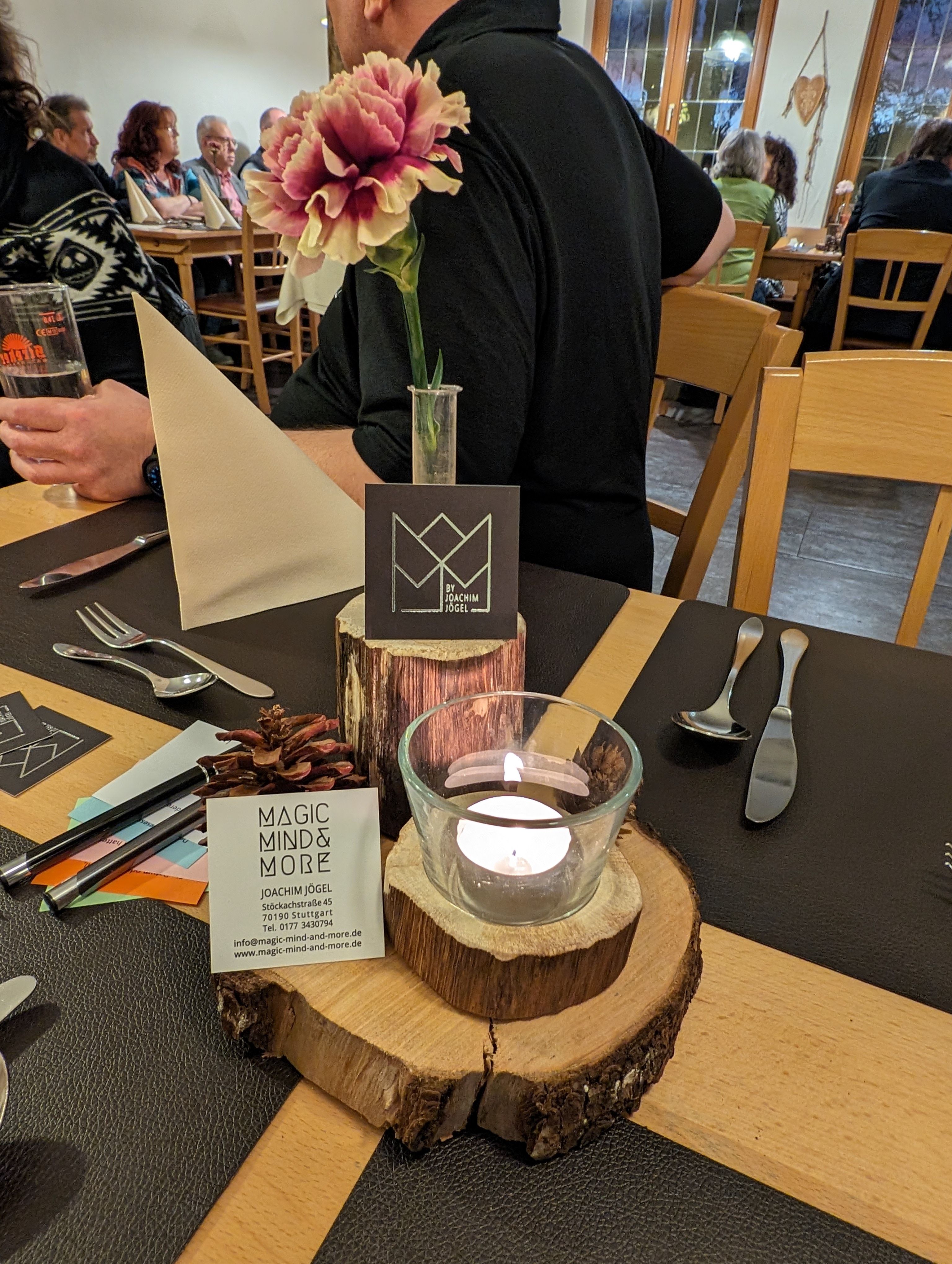 Tischdekoration mit Magic-Mind-&-More-Visitenkarte, Kerze und Blume, im Hintergrund Gäste im Restaurant
